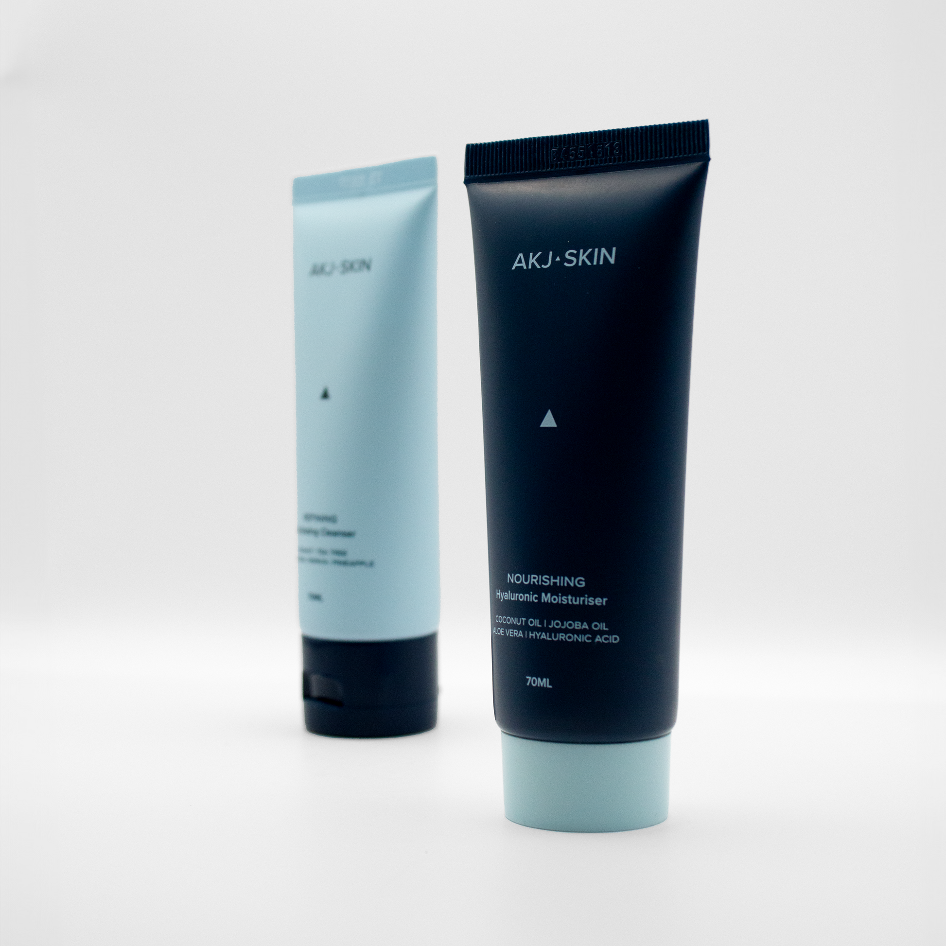 Close-up of AKJ SKIN Nourishing Hyaluronic Moisturiser in focus, with AKJ SKIN Exfoliating Cleanser blurred in the background against a clean white backdrop.